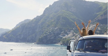 Beautiful happy tourist girl friends enjoying scenic view arms raised of Amalfi Coast on summer road trip adventure vacation in vintage van