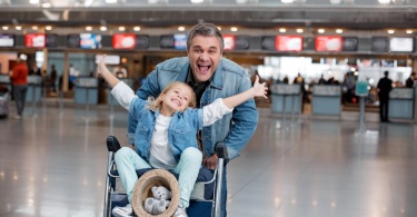 Portrait of joyful mature man is pushing airport trolley with suitcases and his nice little daughter who is sitting on it while rising her hands up