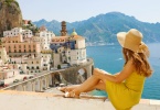 Beautiful young woman with hat sitting on wall looking at stunning panoramic village of Atrani on Amalfi Coast, Italy