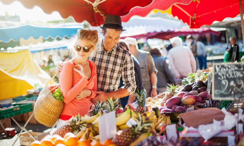 a young couple buying fruits and vegetables in a market on a sunny morning, the young woman carries a basket