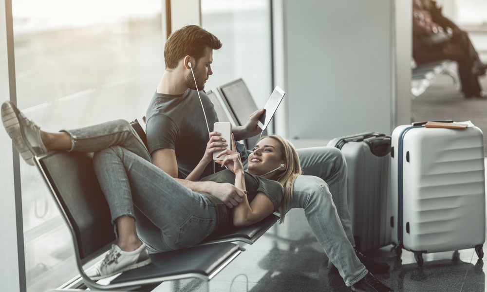 7 Thanksgiving Travel Tips to Make Your Holiday Stress-Free: Full length beaming woman looking at mobile while lying on male legs. He reading book. Satisfied couple locating in airport concept